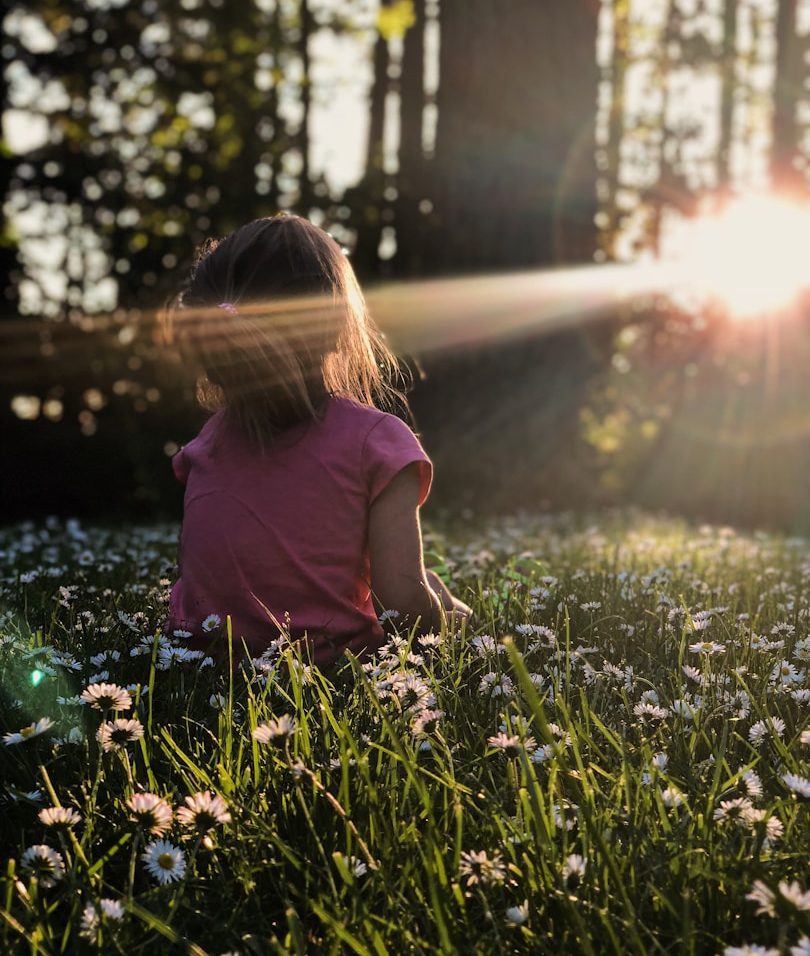 Kind sitzt im Wald in einer Blumenwiese und schaut zur Sonne