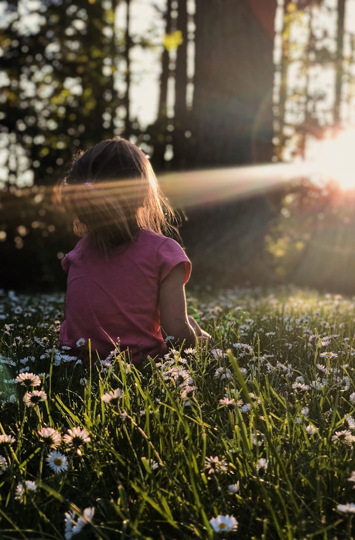 Kind sitzt im Wald in einer Blumenwiese und schaut zur Sonne